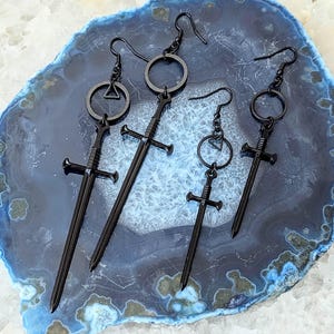 Puede incluir: Cuatro pares de pendientes de espada negros con diferentes estilos de aros. Las espadas están detalladas con una guarda cruzada y una punta puntiaguda. Los pendientes se muestran en una piedra de ágata azul y blanca.