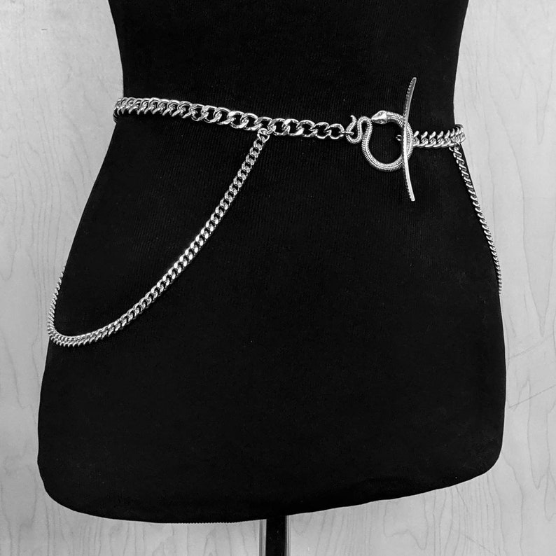 May include: A silver chain belt with a snake-shaped clasp. The chain is thick and has a curb link design. The belt is worn on a black mannequin.