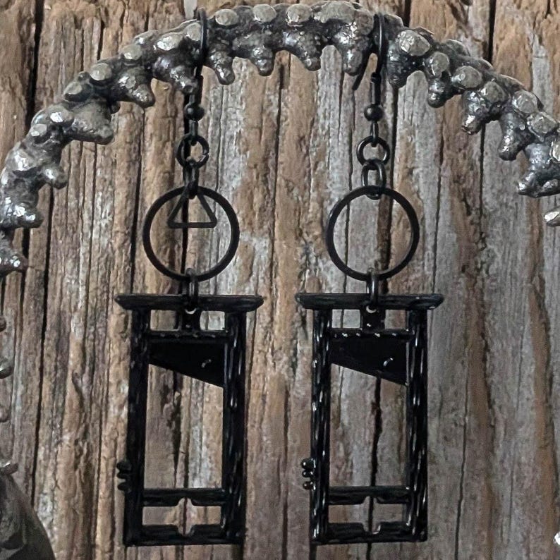 May include: Two black guillotine earrings hanging from a decorative metal arch. Each earring features a guillotine design with a blade and frame. The earrings are suspended from a chain and a circular ring. The arch is textured and has a dark, metallic finish, set against a wooden background.