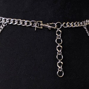 May include: Silver chain belt with a silver clasp and a dangling chain. The chain is attached to a black fabric background.