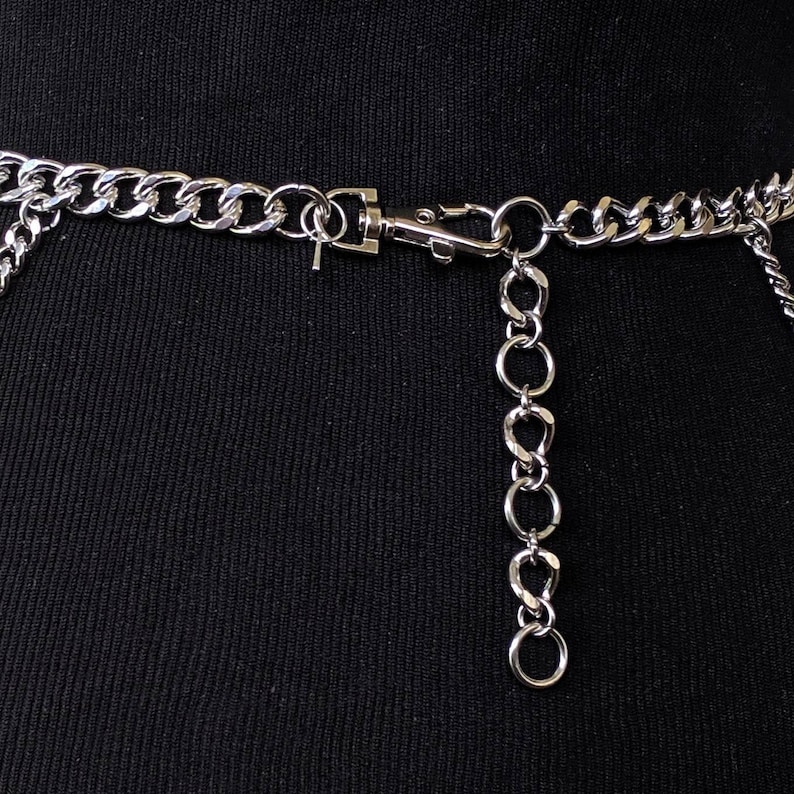 May include: Silver chain belt with a silver clasp and a dangling chain. The chain is attached to a black fabric background.
