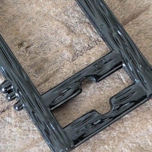 May include: Close-up of a black, rectangular frame with a textured, wood-like appearance. The frame has a glossy finish and features rounded edges and a unique cutout design. The frame is likely for a picture or artwork.