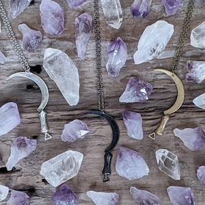 Puede incluir: Tres collares de plata, negro y oro en forma de media luna con un diseño de hoja de hoz. Los collares se muestran sobre una superficie de madera con cristales de cuarzo transparente y amatista.