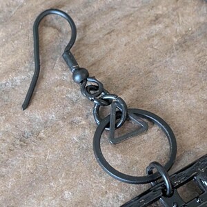 May include: Close-up of a black metal earring with a hook, a small ball, and a geometric design. The earring features a circle with a triangle inside. The metal has a matte finish.