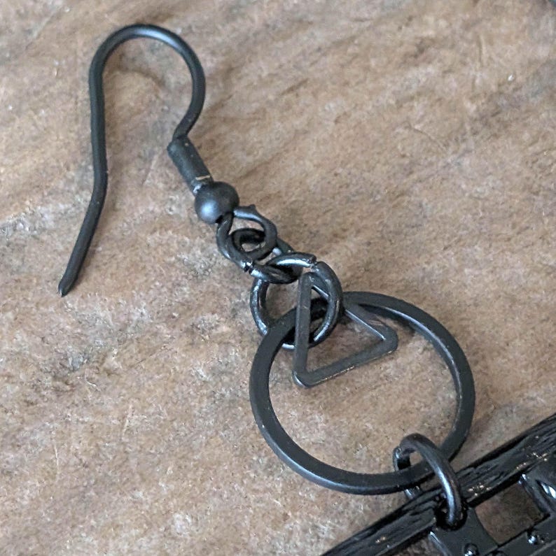 May include: Close-up of a black metal earring with a hook, a small ball, and a geometric design. The earring features a circle with a triangle inside. The metal has a matte finish.