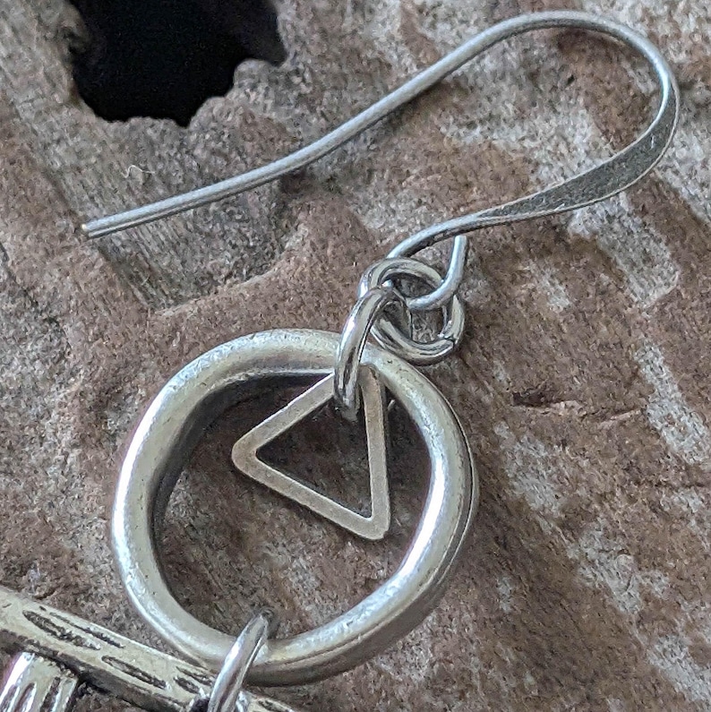 May include: Close-up of a silver earring with a circular design and a triangle inside. The earring has a hook for easy wear. The jewelry is set against a textured, wooden background.