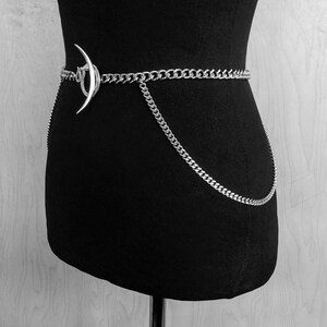 May include: A silver chain belt with a crescent moon charm. The chain is thick and has a curb link design. The belt is worn on a black mannequin.