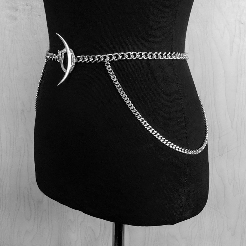 May include: A silver chain belt with a crescent moon charm. The chain is thick and has a curb link design. The belt is worn on a black mannequin.
