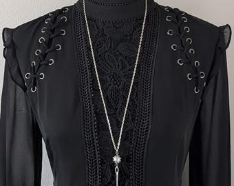 May include: A long silver necklace with a starburst pendant and a dagger charm hangs over a black dress. The dress features lace detailing, eyelet accents, and long sleeves. The overall aesthetic is gothic and elegant.