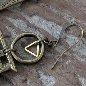 May include: Close-up of a brass earring with a geometric design. The earring features a circular element with a small gold triangle in the center, connected to a hook. The metal has an antique finish, and the background is a wooden surface.