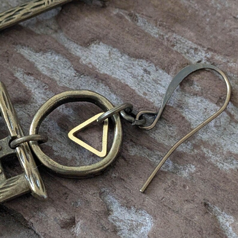 May include: Close-up of a brass earring with a geometric design. The earring features a circular element with a small gold triangle in the center, connected to a hook. The metal has an antique finish, and the background is a wooden surface.