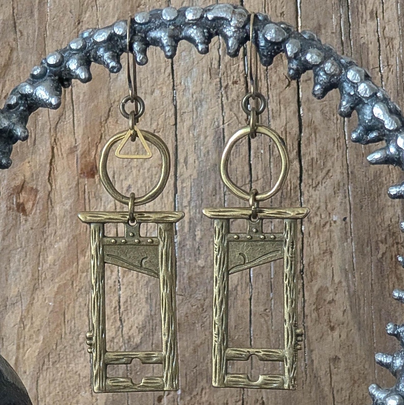 May include: A pair of antique brass-colored earrings. Each earring features a guillotine design suspended from a brass ring and a small triangle. The earrings are displayed against a wooden background and a decorative metal branch.