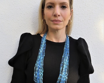 May include: A woman wearing a black top with a long blue and turquoise necklace. The necklace is made of many thin strands of yarn.