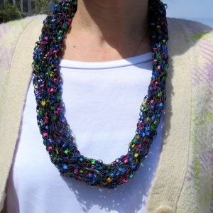 May include: A multi-colored crocheted necklace with blue, green, pink, and gold accents. The necklace is made of a soft, yarn-like material and has a delicate, feminine look.