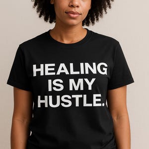 May include: Black crew neck t-shirt with the white text "HEALING IS MY HUSTLE." The shirt is worn by a person with dark curly hair. The background is a solid, neutral color.
