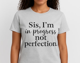 May include: A light gray t-shirt with the text "Sis, I'm in progress not perfection" in black script. The shirt is short-sleeved and worn by a person with dark curly hair and blue jeans.