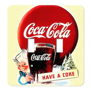 May include: A light switch cover featuring a vintage Coca-Cola advertisement. The design includes a large red circle with the Coca-Cola logo, a glass of Coke, a smiling character, and the text "HAVE A COKE".