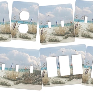 May include: A set of light switch and outlet covers featuring a beach scene with blue water, white sand, and tall grasses. The covers are decorated with a painting of a bird standing in the grass.