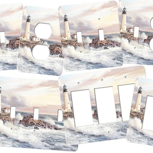 May include: A set of light switch and outlet covers featuring a watercolor painting of a lighthouse on a rocky coast with crashing waves. The lighthouse is white with a black top and is surrounded by blue water and white foam.