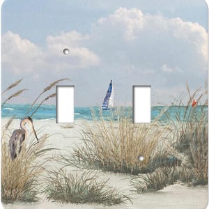 May include: A double light switch wall plate featuring a coastal scene with a heron standing in beach grass, a sailboat on the blue ocean, and a cloudy sky. The rectangular plate has two vertical cutouts and two screw holes.
