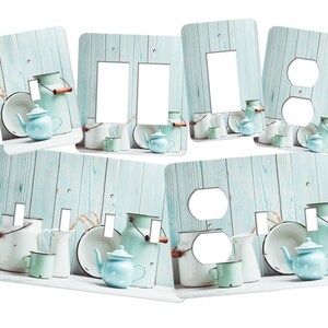 May include: Light blue and white kitchen themed light switch and outlet covers with a rustic wood background. The covers feature a variety of kitchen items, including teapots, pitchers, and bowls.