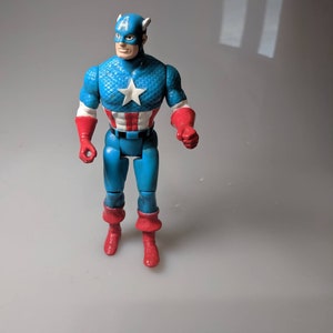 May include: A blue and red plastic action figure of Captain America. The figure has a red, white, and blue costume with a star on the chest. The figure is standing on a white surface.