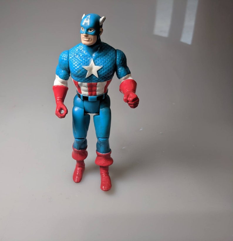 May include: A blue and red plastic action figure of Captain America. The figure has a red, white, and blue costume with a star on the chest. The figure is standing on a white surface.