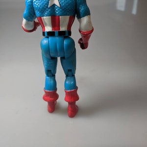 May include: A red, white and blue Captain America action figure with a blue and red suit. The figure is standing with its back to the camera. The figure has a red, white and blue star on its chest.