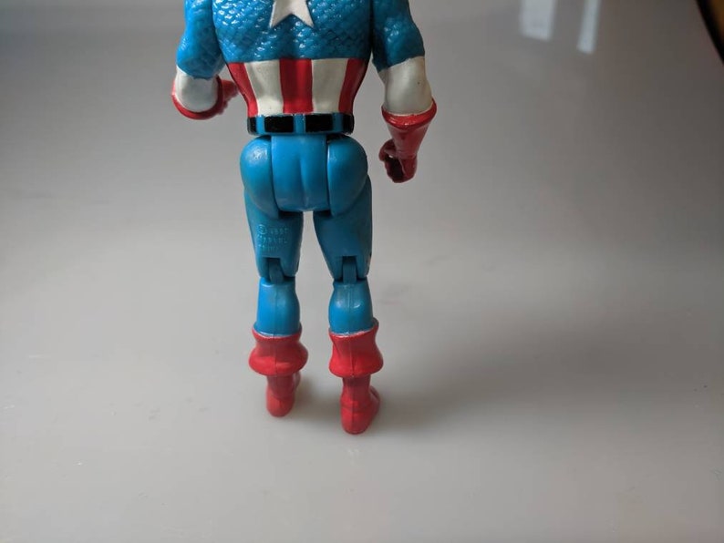 May include: A red, white and blue Captain America action figure with a blue and red suit. The figure is standing with its back to the camera. The figure has a red, white and blue star on its chest.