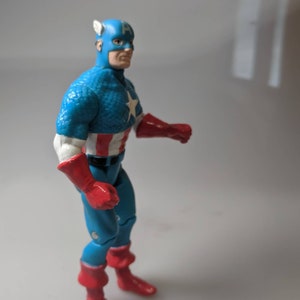 May include: A blue and red Captain America action figure with a white star on his chest. The figure is posed with one arm raised and the other bent at the elbow.