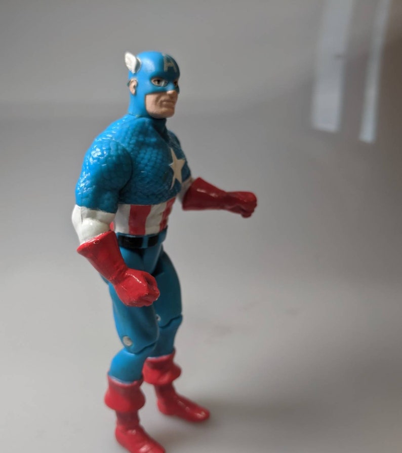 May include: A blue and red Captain America action figure with a white star on his chest. The figure is posed with one arm raised and the other bent at the elbow.