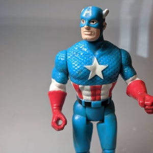 May include: A blue and red plastic action figure of Captain America. The figure has a blue and white star-spangled chest, a red, white, and blue belt, and red gloves. The figure is wearing a blue helmet with a white "A" on the front.