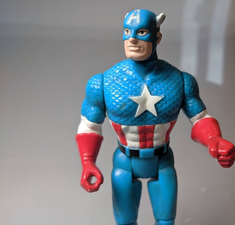 May include: A blue and red plastic action figure of Captain America. The figure has a blue and white star-spangled chest, a red, white, and blue belt, and red gloves. The figure is wearing a blue helmet with a white "A" on the front.