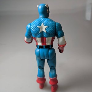 May include: A blue and red plastic action figure of Captain America. The figure is wearing a blue and red costume with a white star on the chest. The figure is standing with its back to the camera.
