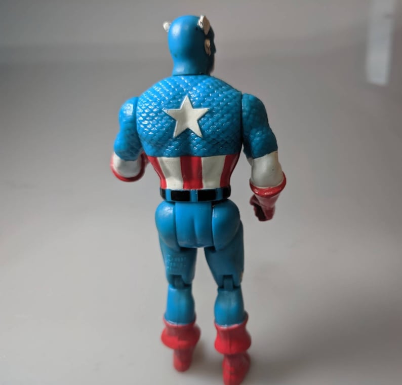 May include: A blue and red plastic action figure of Captain America. The figure is wearing a blue and red costume with a white star on the chest. The figure is standing with its back to the camera.