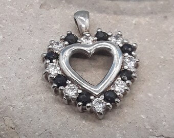 Puede incluir: Un colgante de plata en forma de corazón con un centro transparente y un borde de piedras negras y blancas.