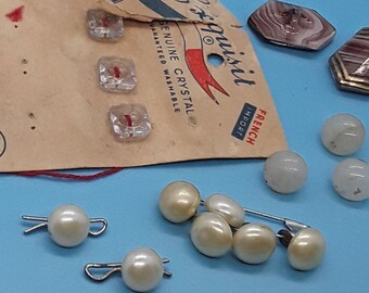 Op de afbeelding: Een assortiment vintage knopen in verschillende kleuren, vormen en maten, waaronder helder glas, bruin, wit en roze. Sommige knopen bevinden zich op kaarten met de tekst "JHN International Buttons" en "Costumakers Guaranteed Washable".