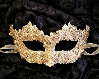 May include: A gold masquerade mask with intricate beading and a delicate lace design. The mask is set against a black lace background.