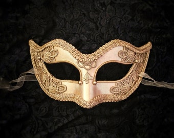 May include: A gold masquerade mask with intricate lace detailing. The mask has a traditional Venetian style with a pointed top and a curved bottom.
