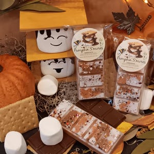 May include: An arrangement of fall-themed treats, including graham crackers, chocolate bars, and marshmallows. Packages of Pumpkin S'mores wax melts are also visible, along with decorative elements like leaves and a small wooden structure with smiling marshmallow faces.