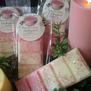 May include: Three wax melts in clear packaging with pink and white glitter. The wax melts are labelled "Bee Fruitty Pink Grapefruit" and feature a bee and grapefruit illustration. The text "BeeWicked Wax Works" is also on the packaging.