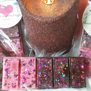 May include: Two wax melts, one pink with glitter and the text "Be Mine Raspberry Drizzle", the other brown with glitter. The wax melts are next to a brown glitter candle.