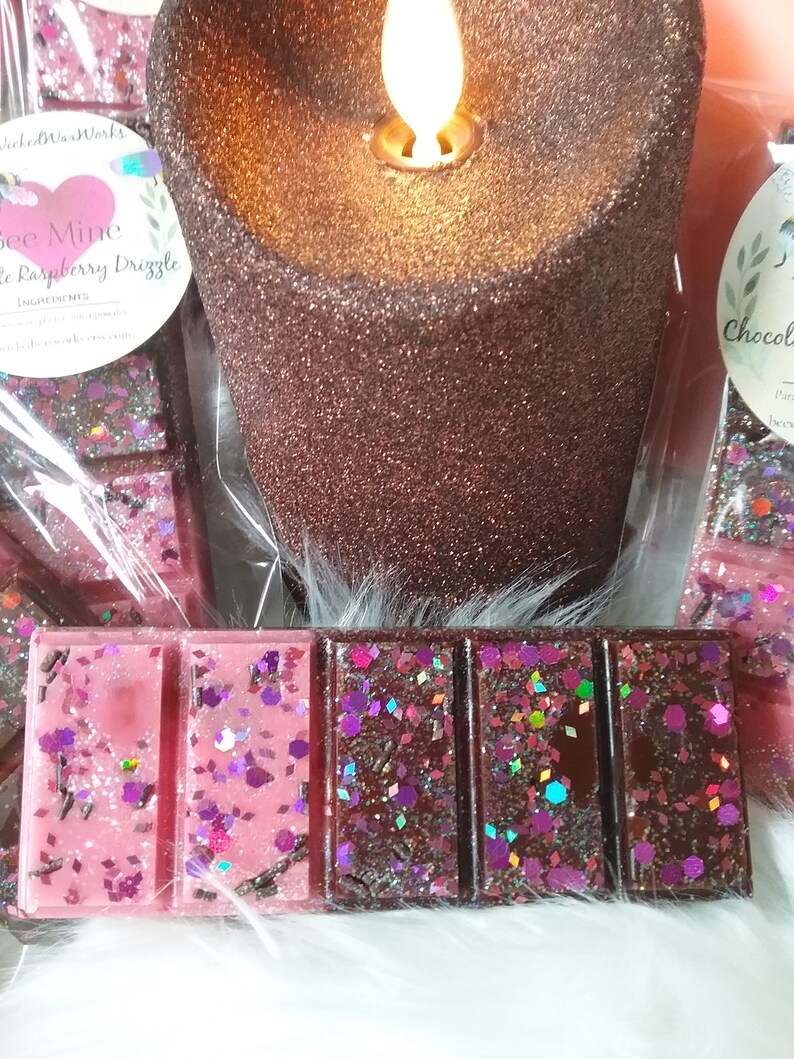 May include: Two wax melts, one pink with glitter and the text "Be Mine Raspberry Drizzle", the other brown with glitter. The wax melts are next to a brown glitter candle.