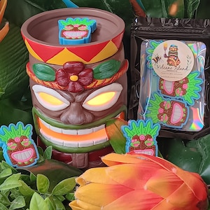 May include: A brown, red, and yellow tiki-themed decorative item with a smiling face and glowing eyes. The image also includes several matching tiki-shaped stickers and a colorful flower arrangement, all set against a backdrop of tropical foliage.