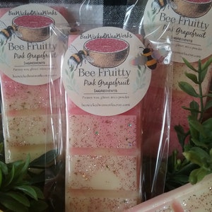 May include: Three wax melts in clear packaging with pink grapefruit scent. The wax melts are layered with pink and white wax and glitter. The packaging has a label that says "Bee Fruitty Pink Grapefruit" and lists the ingredients as "parsoy wax, glitter, mica powder".