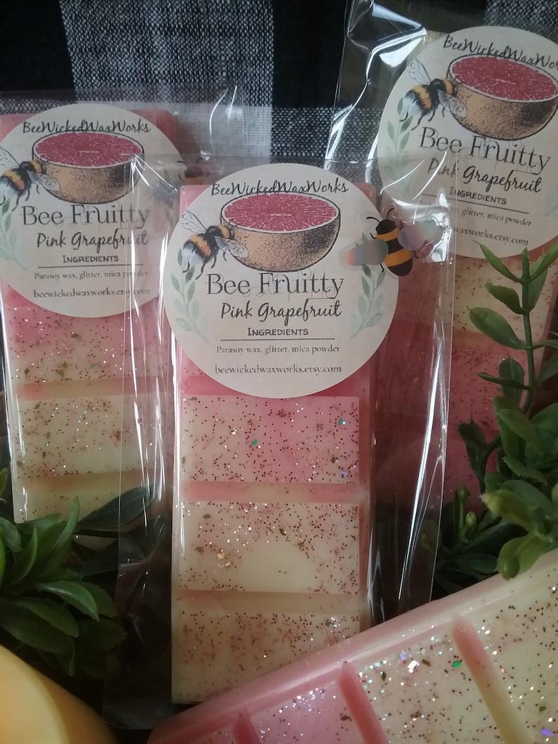 May include: Three wax melts in clear packaging with pink grapefruit scent. The wax melts are layered with pink and white wax and glitter. The packaging has a label that says "Bee Fruitty Pink Grapefruit" and lists the ingredients as "parsoy wax, glitter, mica powder".