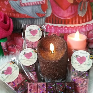May include: Three wax melts in clear packaging with the text "Bee Mine Chocolate Raspberry Drizzle" on the front. The wax melts are pink and brown with glitter. The wax melts are on a white surface with two lit candles and a red and white heart-shaped pillow.