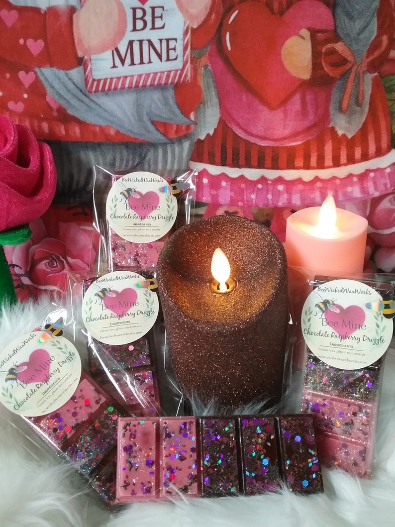 May include: Three wax melts in clear packaging with the text "Bee Mine Chocolate Raspberry Drizzle" on the front. The wax melts are pink and brown with glitter. The wax melts are on a white surface with two lit candles and a red and white heart-shaped pillow.