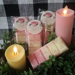 May include: Three pink and white wax melts in clear packaging with a pink grapefruit scent. The packaging has a bee and a watermelon graphic with the text "Bee Fruitty Pink Grapefruit". The wax melts are on a table with two candles, one pink and one yellow.