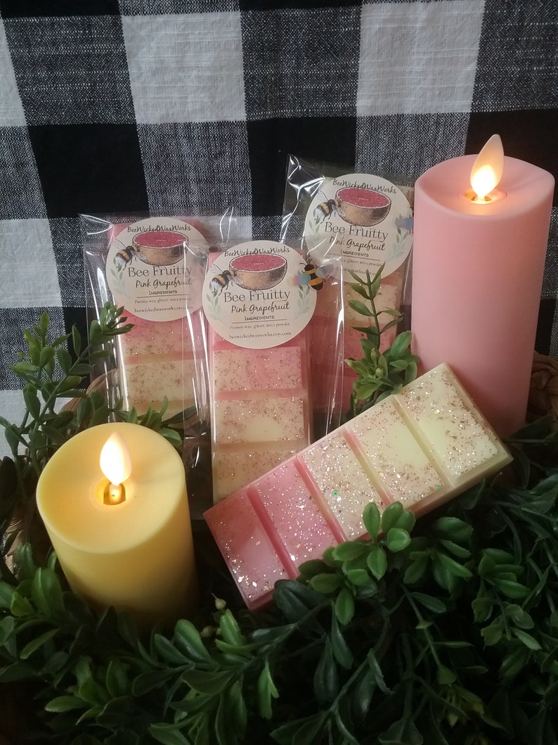 May include: Three pink and white wax melts in clear packaging with a pink grapefruit scent. The packaging has a bee and a watermelon graphic with the text "Bee Fruitty Pink Grapefruit". The wax melts are on a table with two candles, one pink and one yellow.